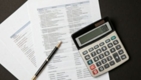 Rounding Reminders Can Prove Costly. Image showcases Calculator over blurred financial documents.